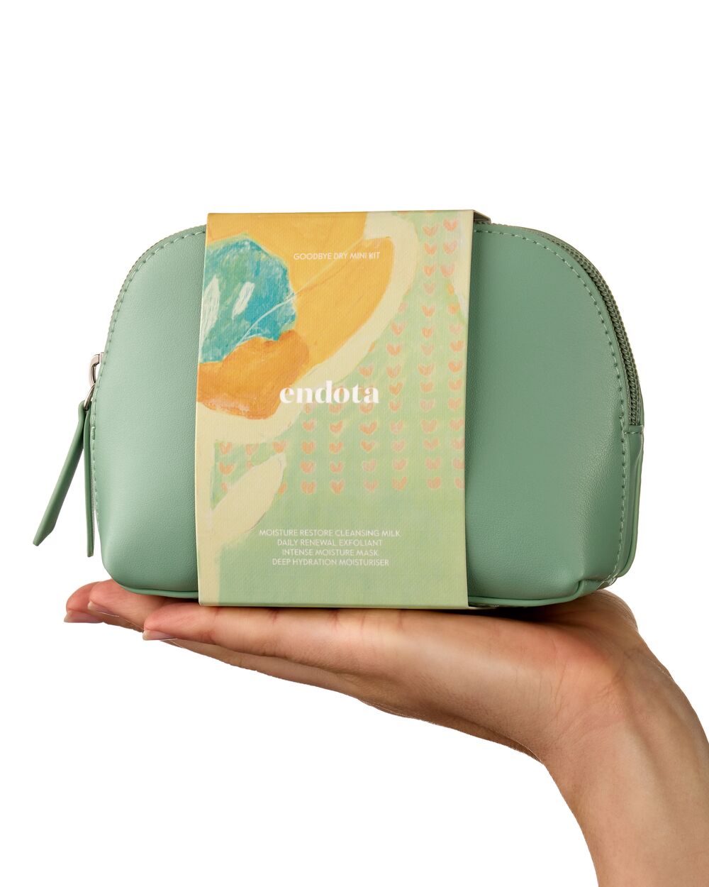 A sage green zippered cosmetic pouch held in a hand against a white background, wrapped in a decorative paper sleeve with the brand name 'endota' and abstract pastel artwork.
