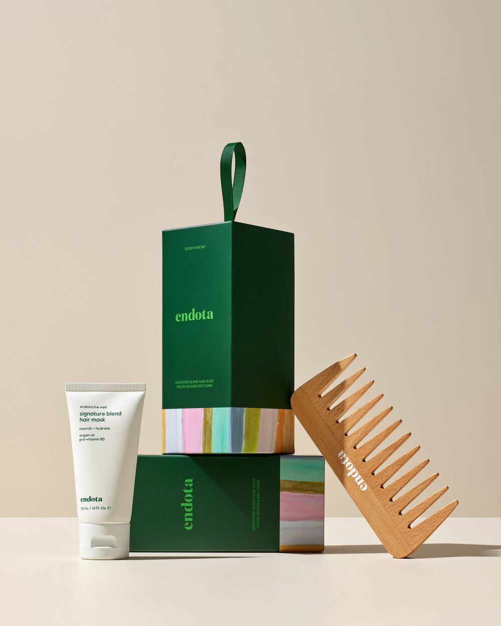 endota haircare gift set featuring a dark green bon bon gift box with a green loop handle, a white tube of signature blend hair mask, and a light wood hair comb, arranged on a beige studio background.