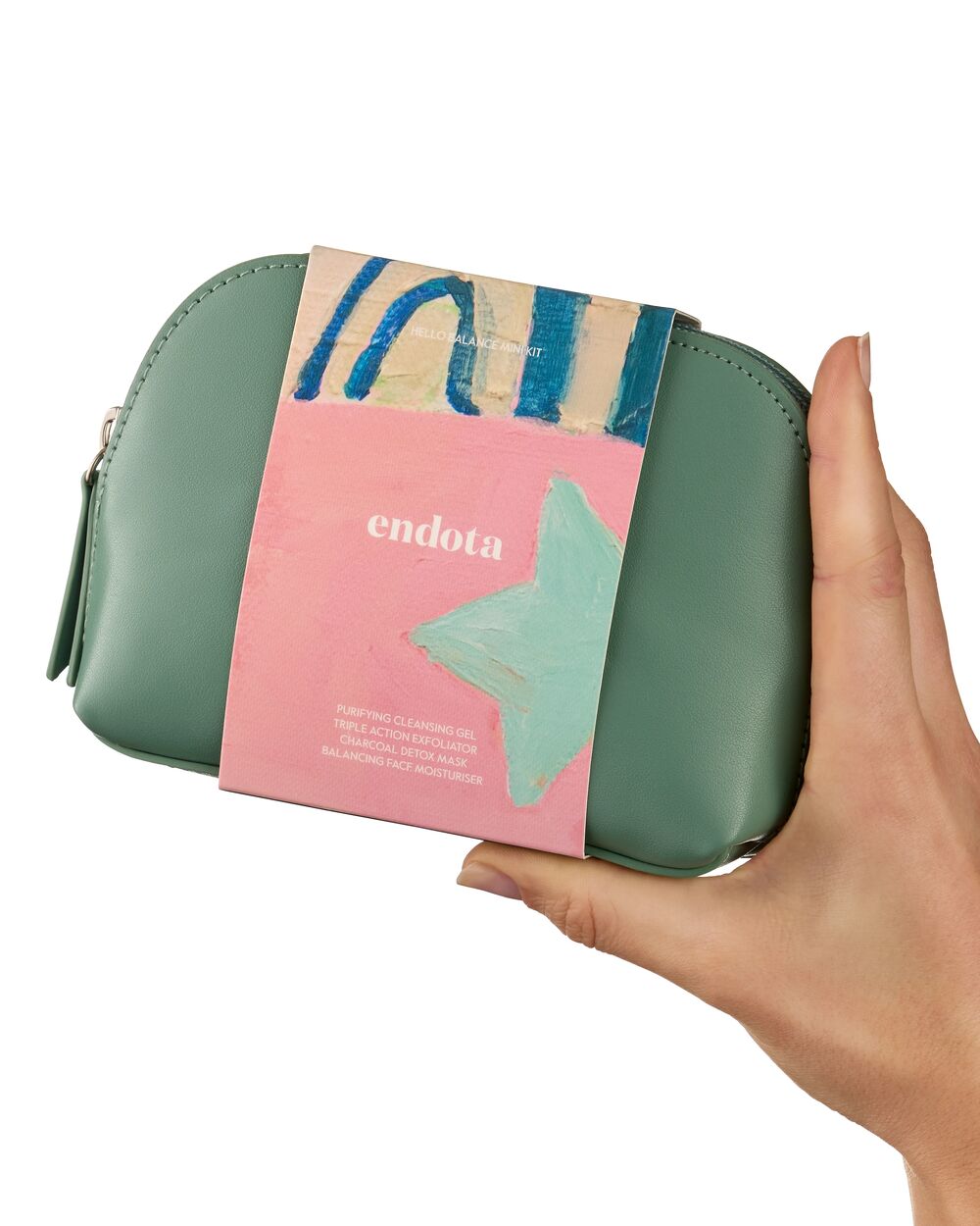 A hand holding a sage green endota skincare pouch with a pink paper sleeve wrapped around it, against a plain white background.