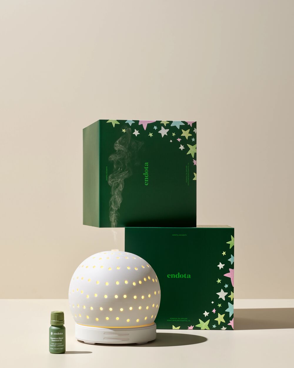 endota diffuser gift set featuring a glowing white spherical diffuser lamp, a small green essential oil bottle, and two dark green branded gift boxes with star patterns, arranged on a beige studio background.