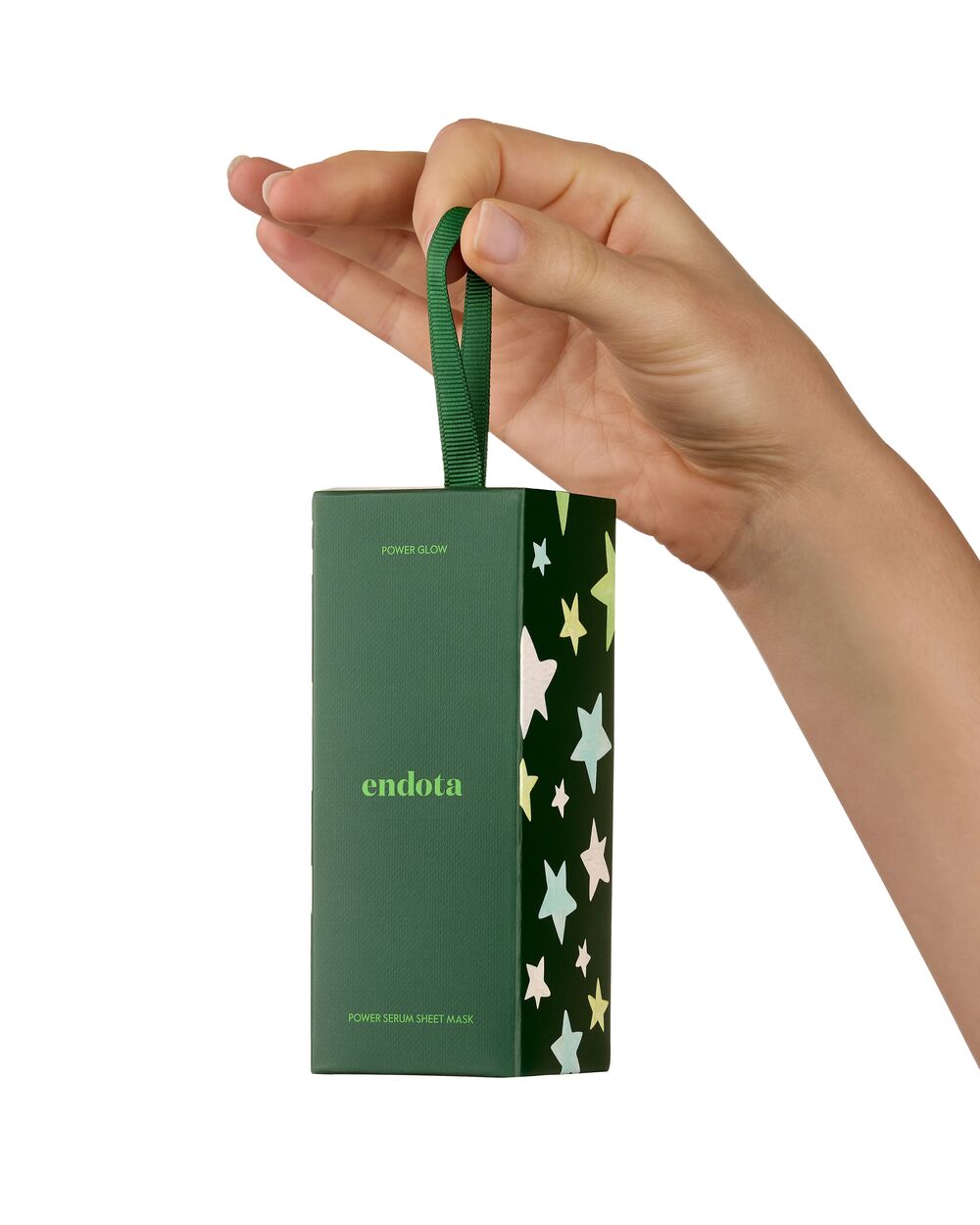 A hand holding a dark green endota skincare box with a matching green ribbon loop handle. The box features the brand name 'endota' in light green on the front, with a side panel decorated with a pattern of light green, yellow, and white stars on a dark green background.