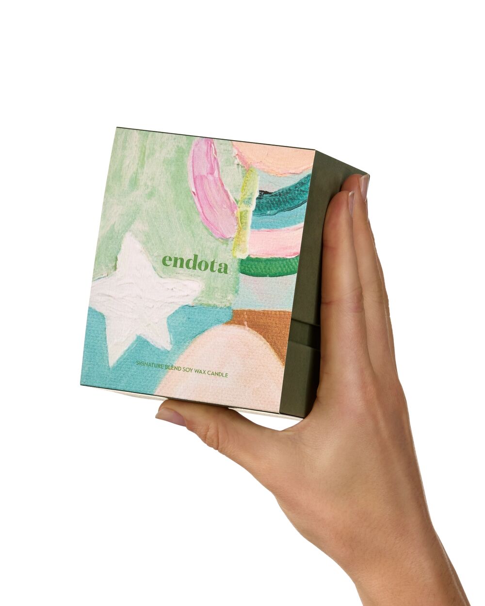 A hand holding a square endota Soy Wax Candle gift box against a solid white background. The box features an abstract, colourful painted design with pastel shades of green, pink, blue, and white, and the brand name 'endota' is printed in green on the front.