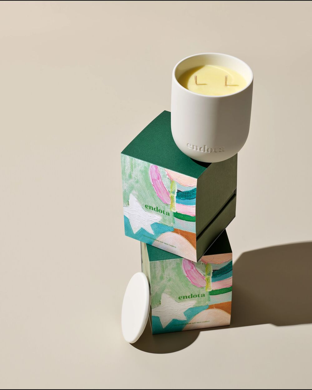 A white endota soy wax candle in a matte white ceramic holder sits atop stacked decorative boxes with a colourful abstract pattern, on a warm beige studio background.
