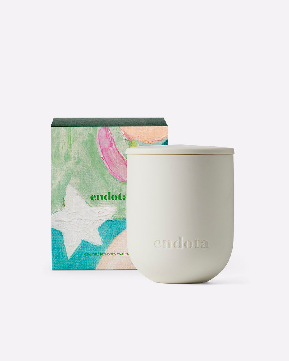 An endota soy wax candle in a matte off-white ceramic jar with a matching lid, shown next to its colorful illustrated product gift box on a clean white background.