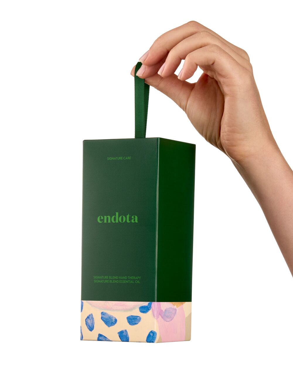 A hand holding a dark green rectangular product gift box with a matching green handle. The box is branded 'endota' and features a light pink and beige patterned lower section with blue accents, set against a plain white background.