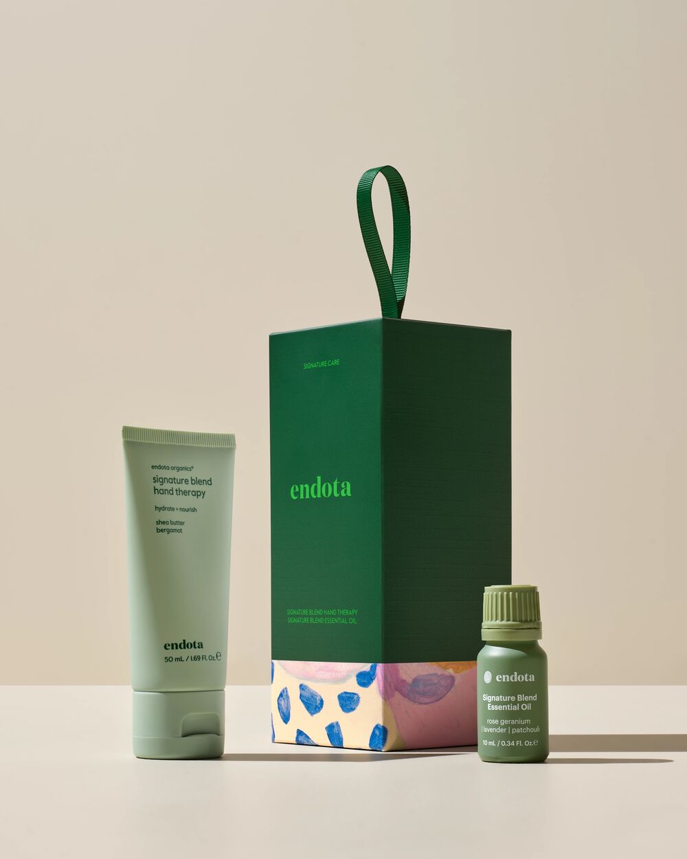 endota skincare gift set featuring a green hand cream tube, a dark green gift box with a colorful patterned base, and a small green essential oil bottle on a beige background.