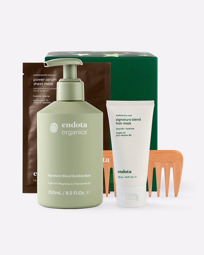 endota Organics gift set featuring a Signature Blend Bubble Bath pump bottle, a white Signature Blend Hair Mask tube, a brown Power Serum Sheet Mask pouch, a wooden comb and a green gift box, arranged on a light grey background.