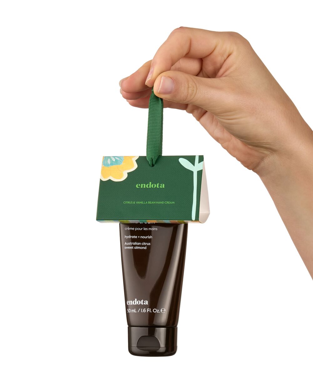 A hand holding a small endota hand cream bottle with its green hanging card packaging against a plain white background.