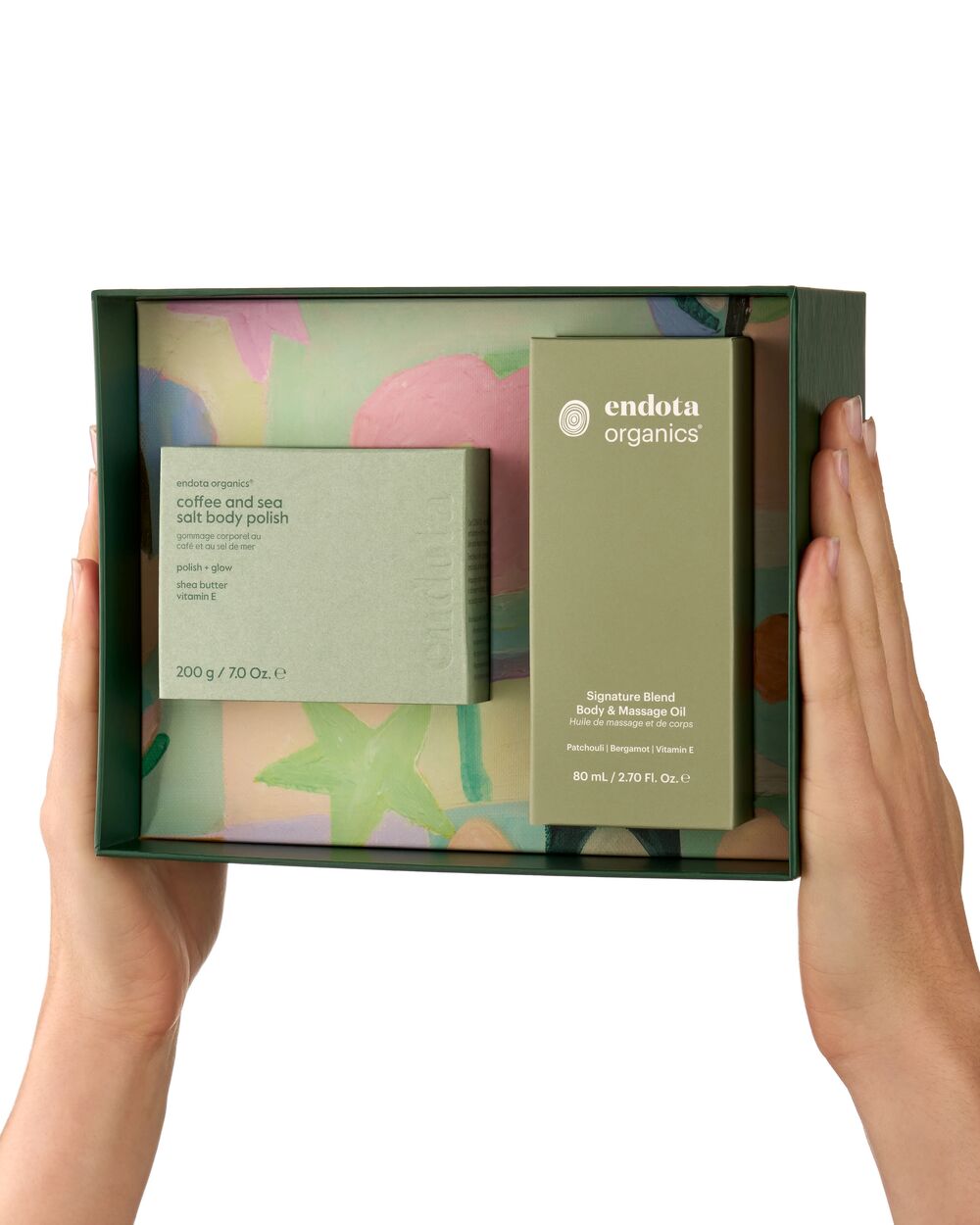 Two hands holding an open dark green gift box containing endota Organics body care products, including a coffee and sea salt body polish and a signature blend body and massage oil, against a plain white background.