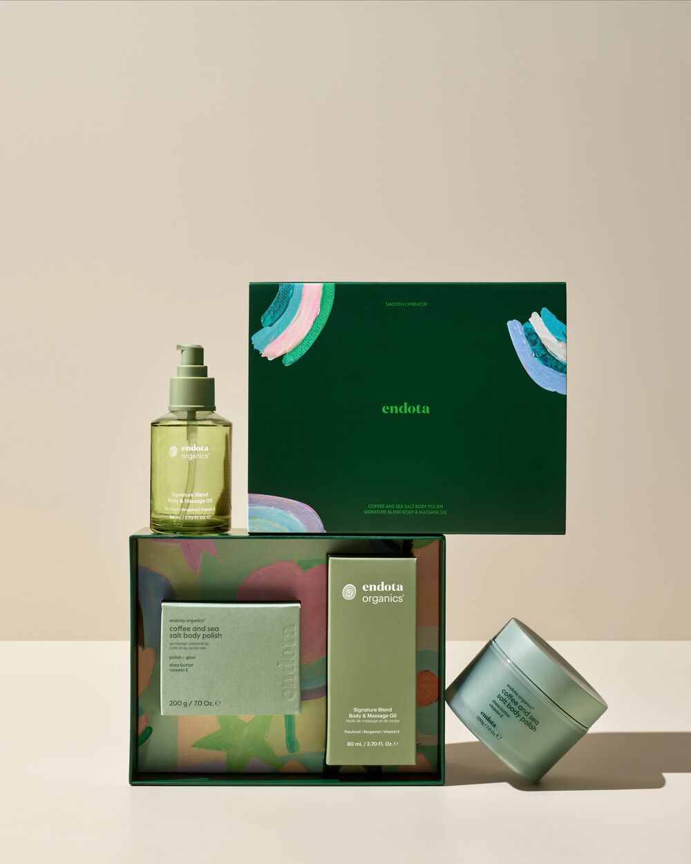 endota Organics body care gift set featuring a green massage oil bottle, a green gift box with colourful abstract accents, a mint green body scrub jar, arranged on a beige studio background.