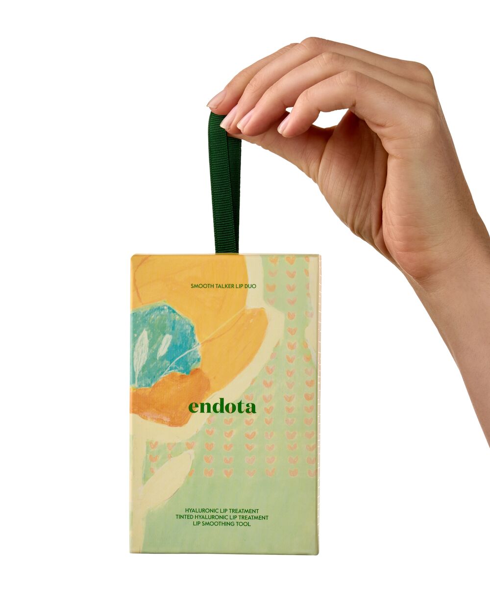 A hand holding a small rectangular product gift box with a dark green loop handle. The box has an abstract pastel design with green, yellow, orange, and blue shapes, and the brand name 'endota' printed in the centre.