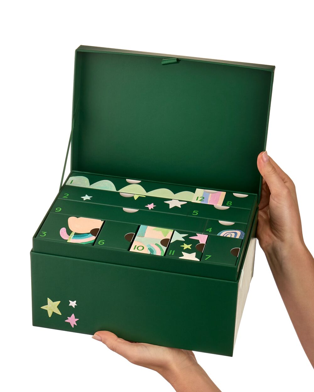 A dark green advent calendar box with multiple illustrated compartments opened to reveal numbered doors, held open by two hands against a plain white background.