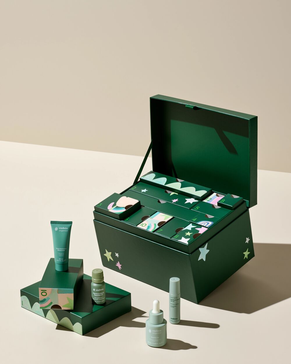 A green skincare gift set displayed on a beige studio background, featuring an open decorative skincare advent calendar box with multiple skincare items inside and a matching green box with a tube and small bottles in front.