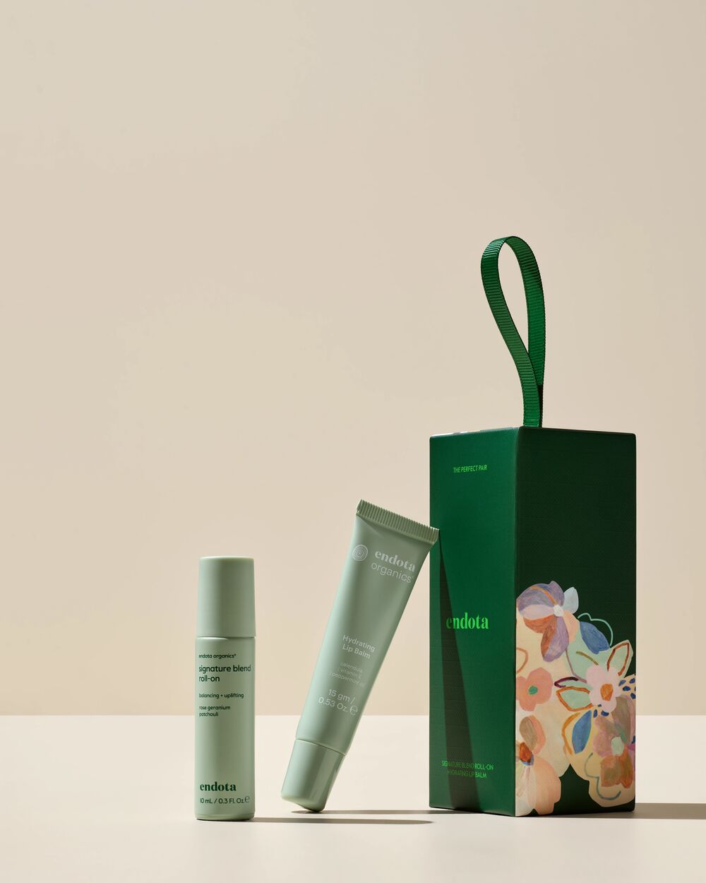 A skincare set from endota on a warm beige background, featuring a light sage green essential oil roll-on, a matching lip balm squeeze tube, and a dark green gift box with a floral illustration and green handle.