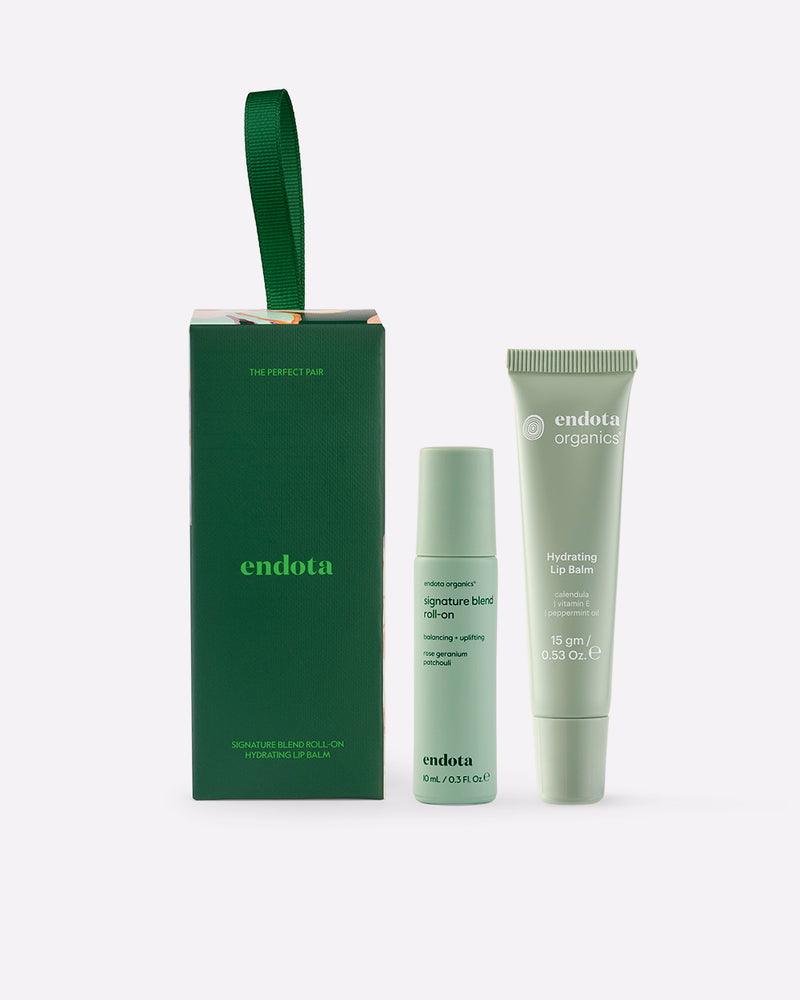 endota gift set featuring a green gift box with a green ribbon handle, a signature blend roll-on bottle, and a hydrating lip balm tube, all in a light sage green color, on a light grey background.